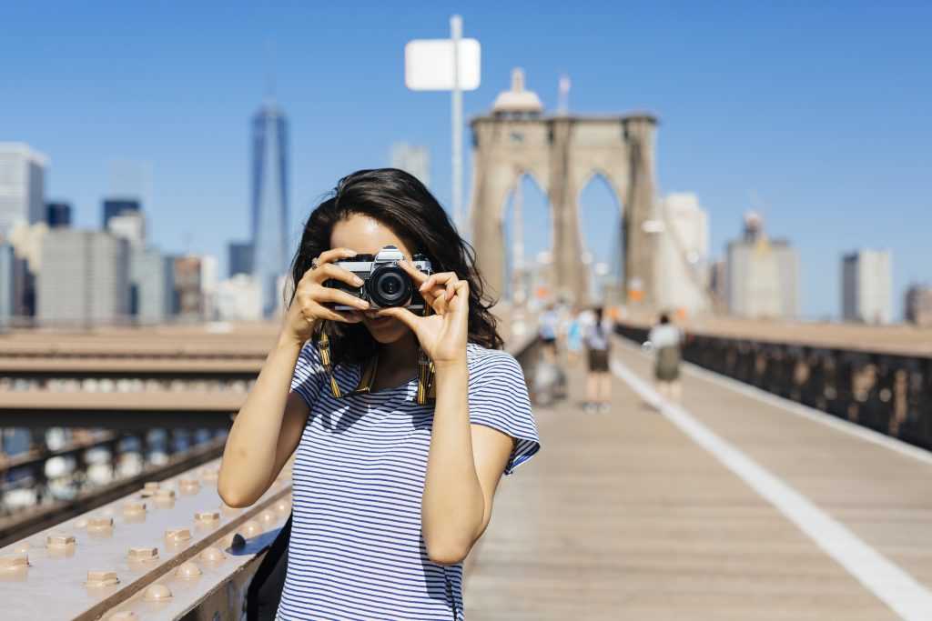 USA, New York City, young woman standing on Brooklyn Bridge taking a photo with camera